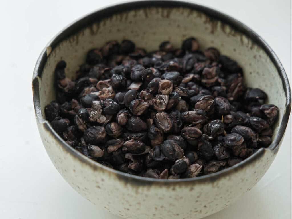 Viral Dense Bean Salad Recipe - Korean BBQ Style. 9 A ceramic bowl filled with black beans rests on a white surface. The beans appear cooked or roasted, and the bowl has a speckled, rustic design.