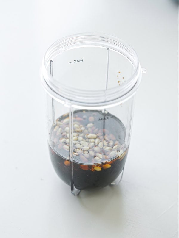 Viral Dense Bean Salad Recipe - Korean BBQ Style. 12 A clear blender cup containing soy sauce and whole peppercorns sits on a light-colored surface, viewed from above.