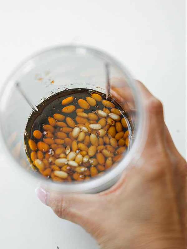 Viral Dense Bean Salad Recipe - Korean BBQ Style. 13 A hand holding a clear cup filled with brown liquid and soaked pine nuts on a white surface.
