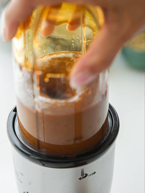 Viral Dense Bean Salad Recipe - Korean BBQ Style. 14 A close-up of a hand holding a small blender with a brown liquid being blended inside a transparent container. The blender base is white and black, and the background is blurred.