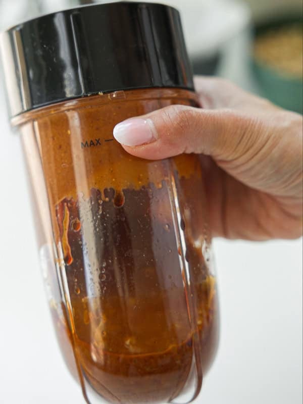 Viral Dense Bean Salad Recipe - Korean BBQ Style. 15 A hand holding a blender cup filled with reddish-brown sauce, showing the liquid level just below the MAX line marked on the cup.