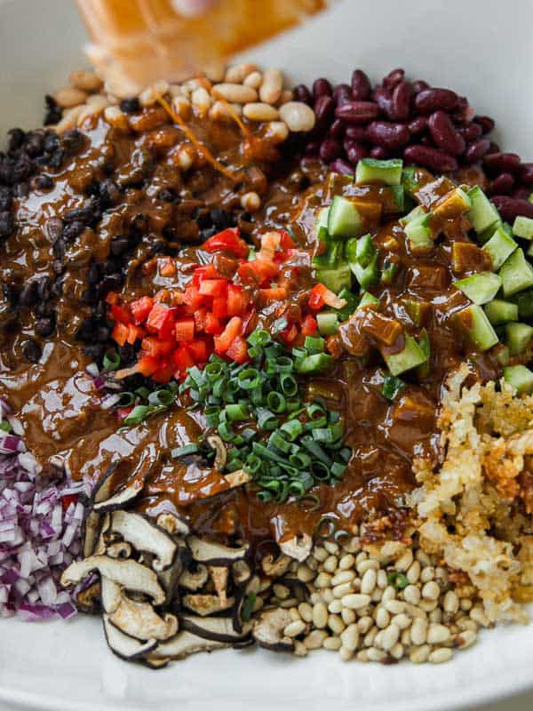 Viral Dense Bean Salad Recipe - Korean BBQ Style. 17 A colorful salad bowl with sections of black beans, kidney beans, white beans, diced red pepper, cucumber, chopped green onions, red onion, grilled eggplant, pine nuts, and crispy rice, topped with brown sauce.