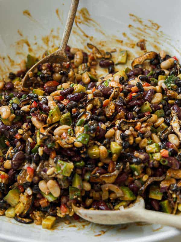 Viral Dense Bean Salad Recipe - Korean BBQ Style. 19 A close-up of a colorful dense bean salad with diced vegetables, black beans, kidney beans, pinto beans, corn, red peppers, and herbs, mixed together in a white bowl with two wooden serving spoons.