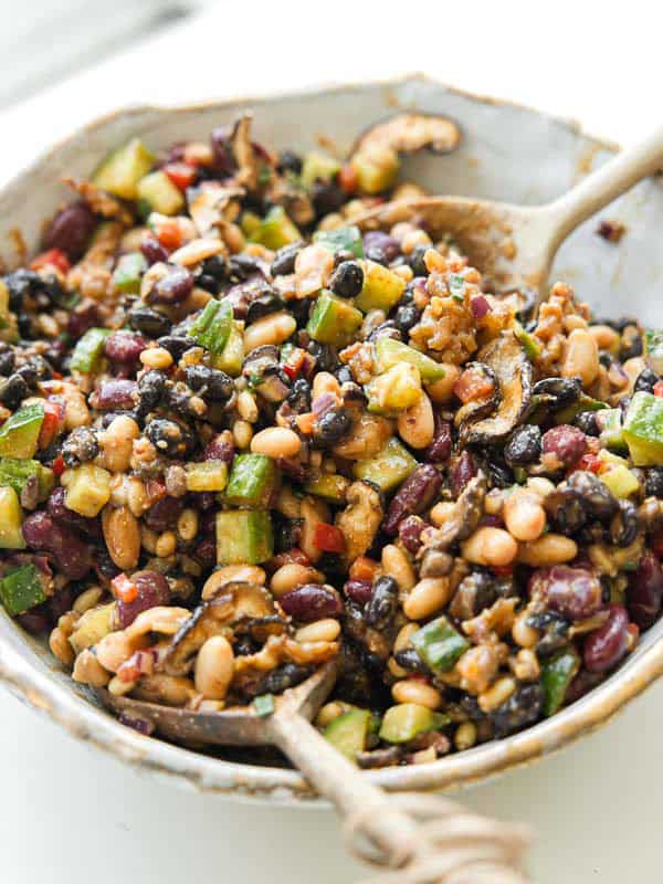 Viral Dense Bean Salad Recipe - Korean BBQ Style. 4 A bowl filled with a colorful dense bean salad containing black beans, kidney beans, navy beans, chopped cucumber, diced red pepper, and mushrooms, mixed together with a Korean BBQ dressing. A spoon rests in the bowl.