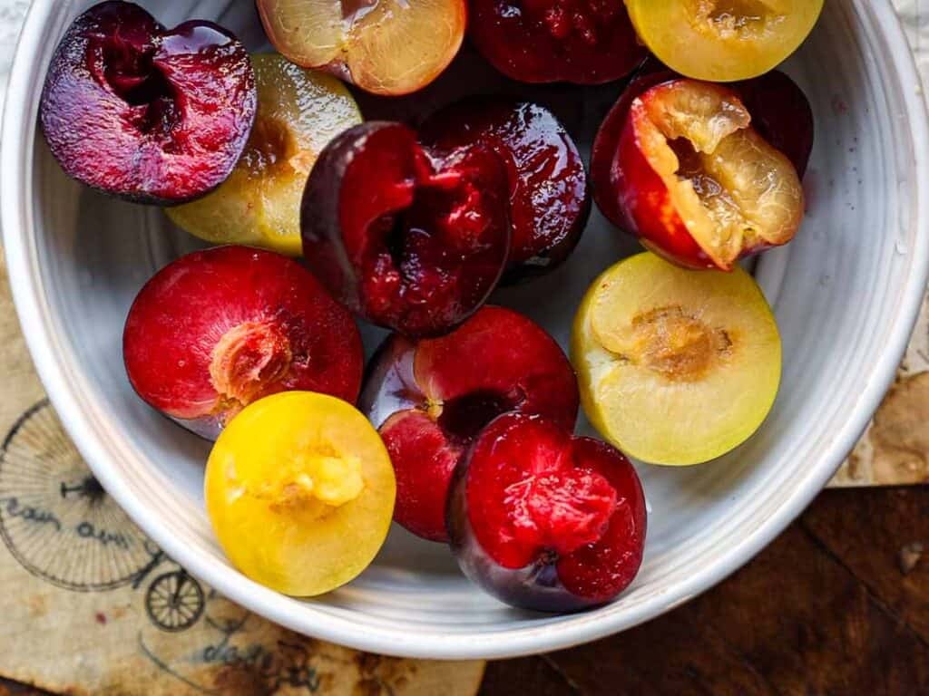 7 Ingredient Vegan Plum Torte - Easy & Delicious. 37 A white bowl filled with halved red and yellow plums, showing their juicy flesh and glossy skins, sits on a table with part of a vintage-style paper visible underneath.