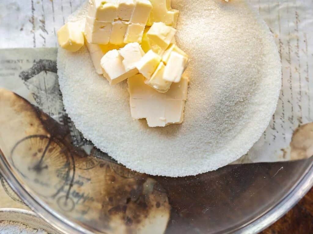 7 Ingredient Vegan Plum Torte - Easy & Delicious. 40 A glass bowl containing granulated sugar and cubed vegan butter sits on a vintage-style paper background.