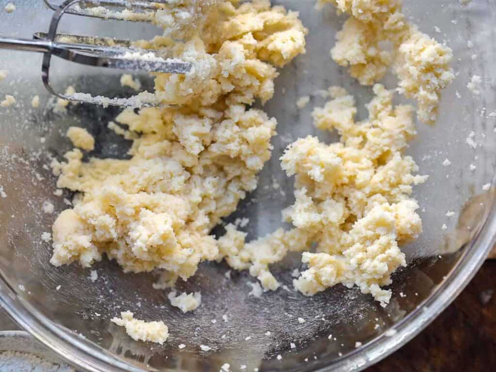 7 Ingredient Vegan Plum Torte - Easy & Delicious. 41 A close-up of a glass bowl with creamed vegan butter and sugar being mixed by a metal hand mixer. The mixture is light yellow and crumbly, with some bits sticking to the sides of the bowl.