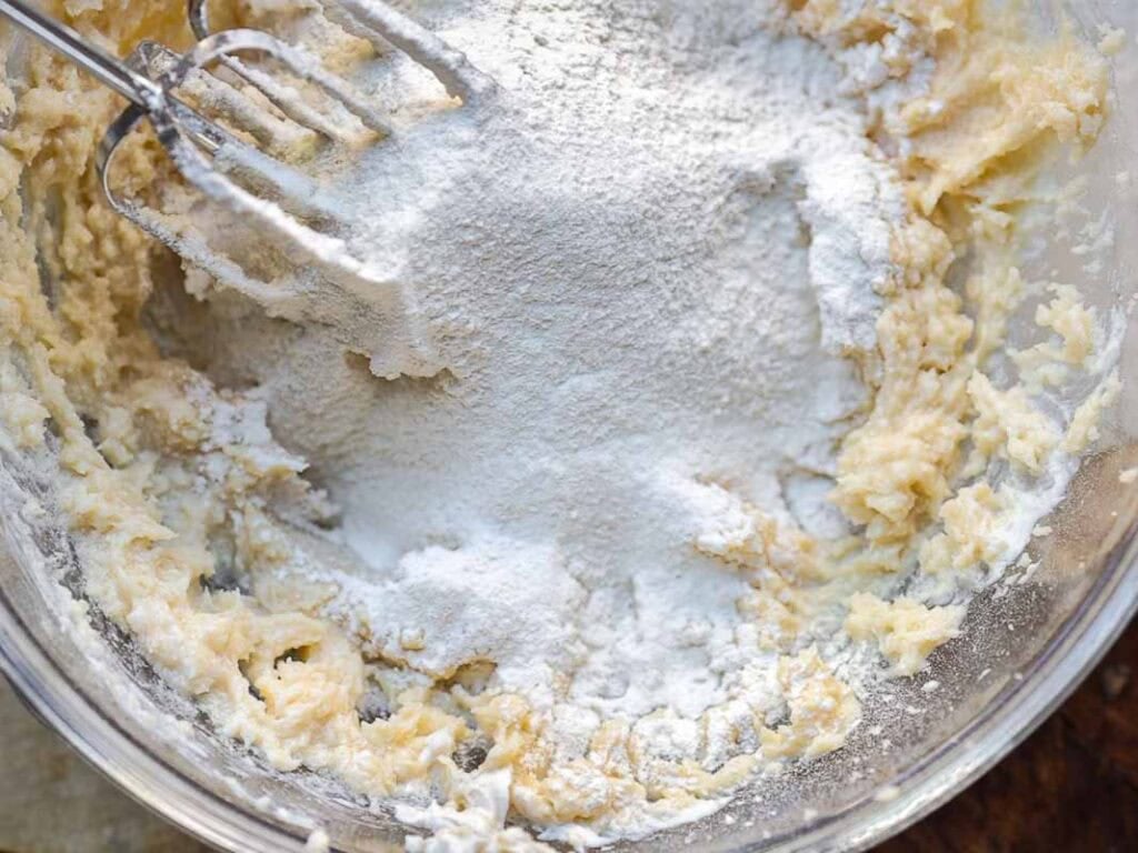 A glass bowl containing creamed butter and sugar, with flour sprinkled on top, and electric mixer beaters partially visible, ready to blend the ingredients together for baking.