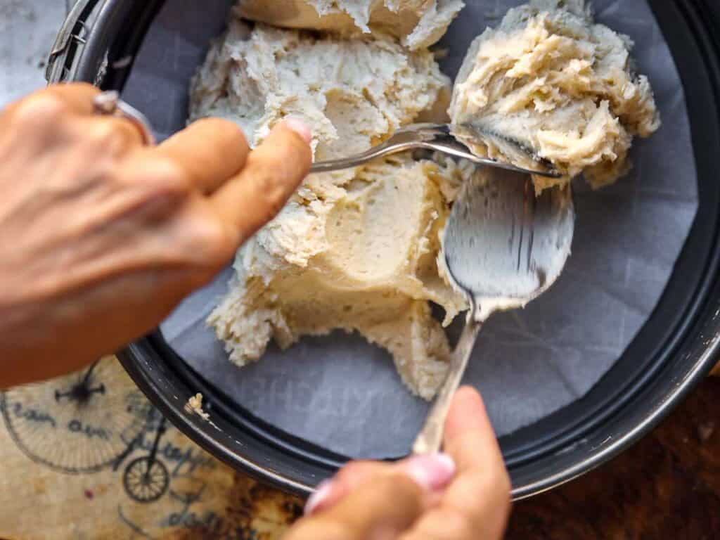 7 Ingredient Vegan Plum Torte - Easy & Delicious. 46 Close-up of hands using two spoons to spread a thick, creamy batter into a round, parchment-lined baking pan. The image suggests preparation for baking, possibly a cake or cheesecake base.