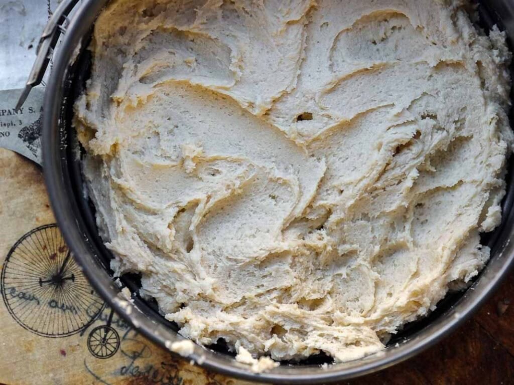 7 Ingredient Vegan Plum Torte - Easy & Delicious. 47 A close-up of a round baking tin filled with thick, creamy cake batter, ready to be baked. The tin sits on a rustic surface with vintage-style designs and text partially visible underneath.