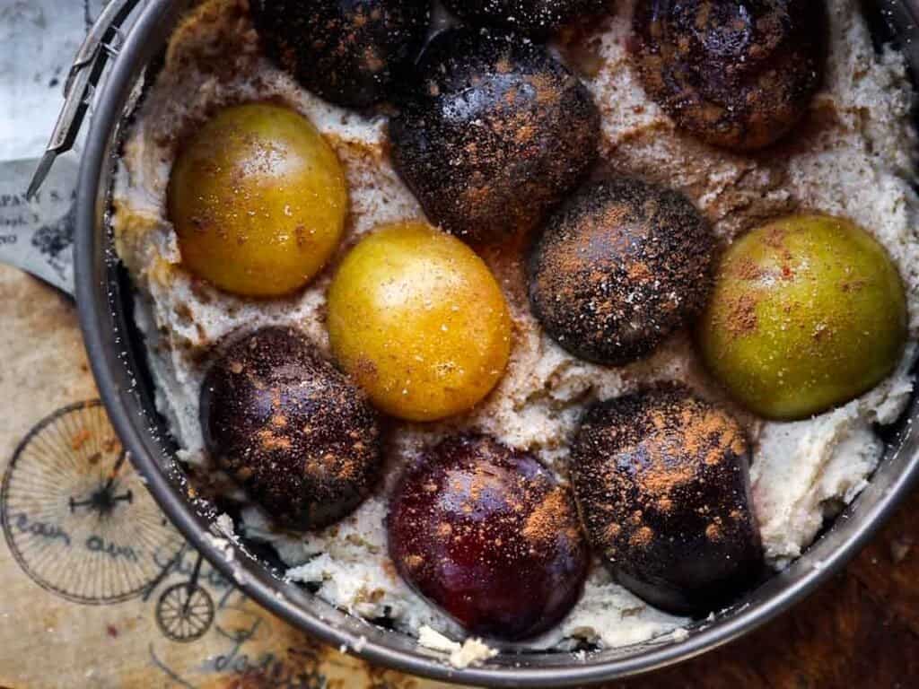 7 Ingredient Vegan Plum Torte - Easy & Delicious. 49 A close-up of a baking pan filled with a creamy batter topped with whole plums of varying colors, dusted with cinnamon, ready to be baked. The pan rests on a brown paper with bicycle illustrations.