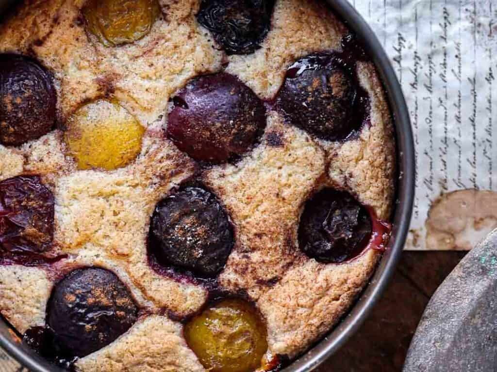 7 Ingredient Vegan Plum Torte - Easy & Delicious. 34 A close-up of a rustic vegan plum torte in a round metal baking pan, featuring whole baked plums nestled in golden-brown, slightly textured cake. Handwritten paper is partially visible beneath the pan.