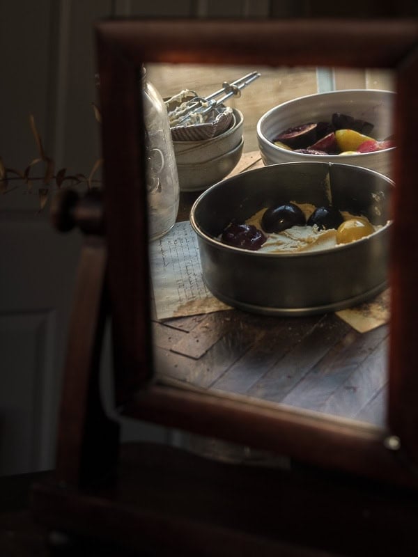 7 Ingredient Vegan Plum Torte - Easy & Delicious. 50 A wooden-framed mirror reflects two metal baking pans filled with colorful ingredients, sitting on a rustic wooden table with a vintage egg beater and glass jar in the background.