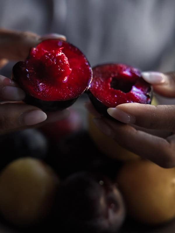 7 Ingredient Vegan Plum Torte - Easy & Delicious. 38 A person gently holds and pulls apart a ripe, juicy red plum, revealing its vibrant, glistening interior. More whole plums and blurred fruit are visible in the background.