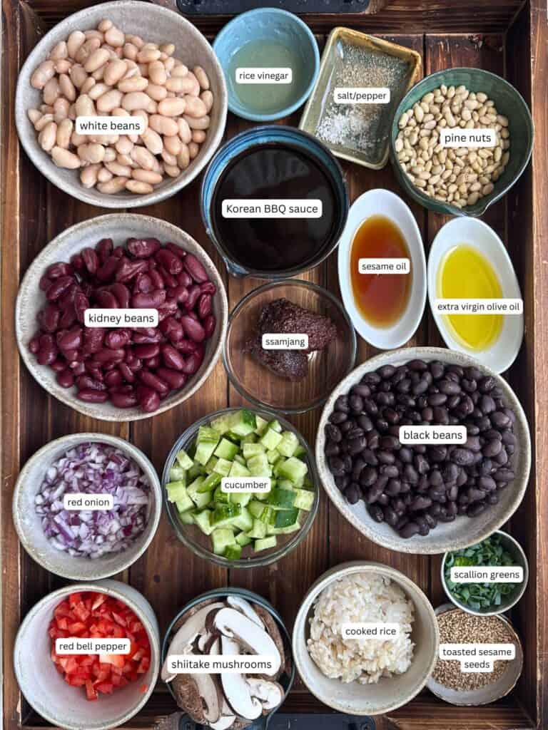 Viral Dense Bean Salad Recipe - Korean BBQ Style. 5 A wooden tray with bowls containing ingredients for dense bean salad: white beans, kidney beans, black beans, red onion, red bell pepper, cucumber, shiitake mushrooms, cooked rice, pine nuts, rice vinegar, salt/pepper, BBQ sauce, ssamjang, oils, scallion greens, and sesame seeds.