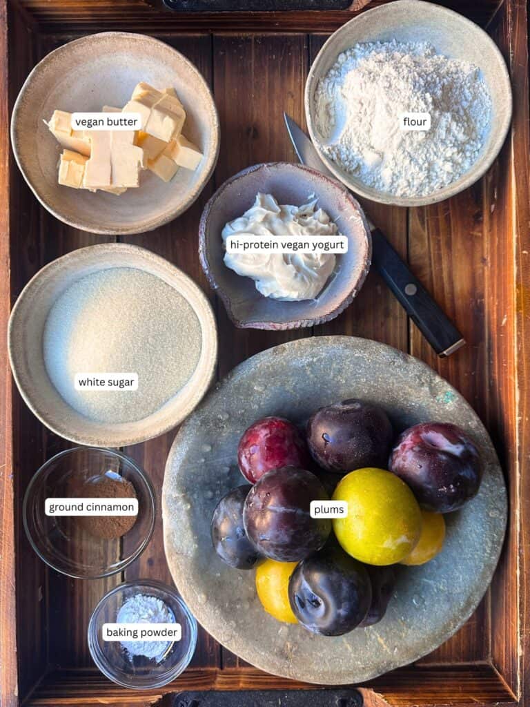 7 Ingredient Vegan Plum Torte - Easy & Delicious. 36 A wooden tray with bowls containing labeled ingredients: vegan butter, flour, white sugar, high-protein vegan yogurt, ground cinnamon, baking powder, and a plate of assorted plums, with a knife beside the yogurt.