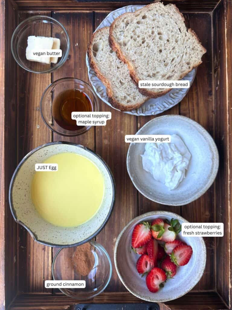 5-Ingredient Vegan French Toast Recipe - Fast & Easy! 4 A wooden tray holds bowls and plates with ingredients for vegan French toast: vegan butter, maple syrup, stale sourdough bread, vegan vanilla yogurt, JUST Egg, strawberries, and ground cinnamon, each labeled.