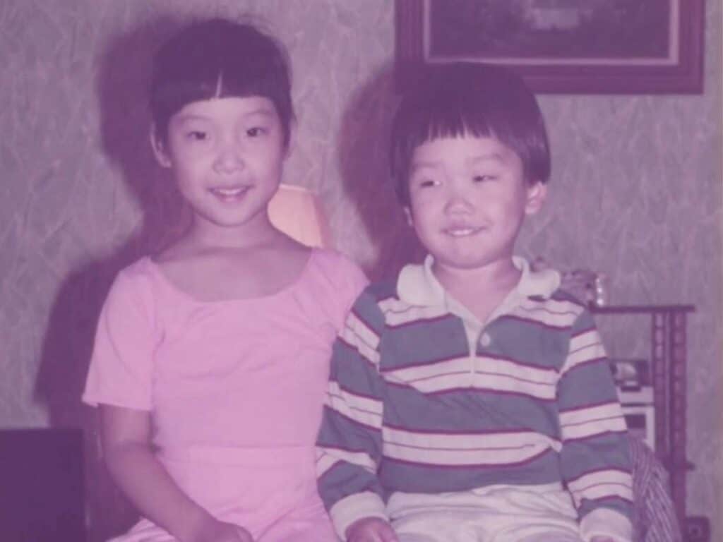 An ice cream truck and my little brother. 1 A young Joanne in a pink dress and her little brother Jaesun, in a striped shirt and light pants, sit side by side on a wooden bench, smiling in a warmly lit room with patterned wallpaper and a framed picture in the background.