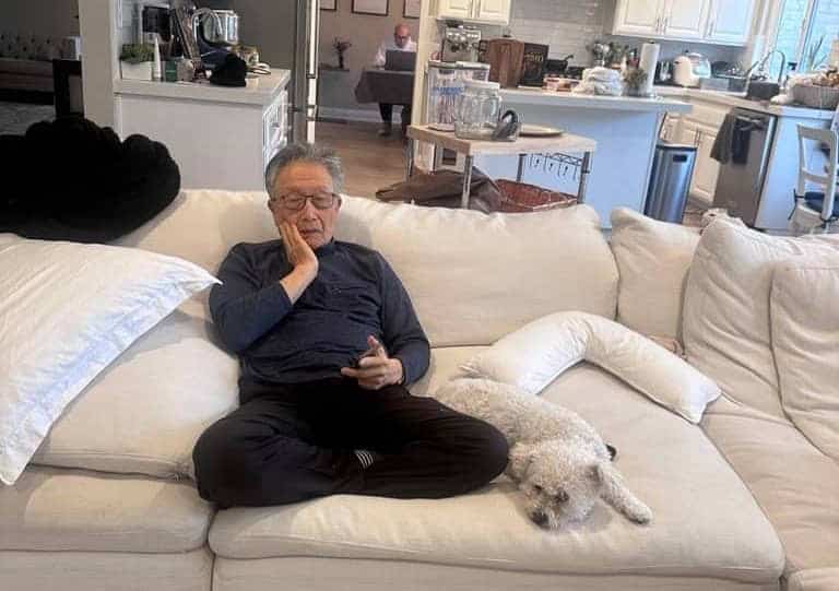 Should influencers get political?? 2 An older man sits cross-legged on a white couch, holding his face while looking at his phone, perhaps scrolling through social media. A small white dog lies next to him. In the background, another person works at a table in the kitchen.