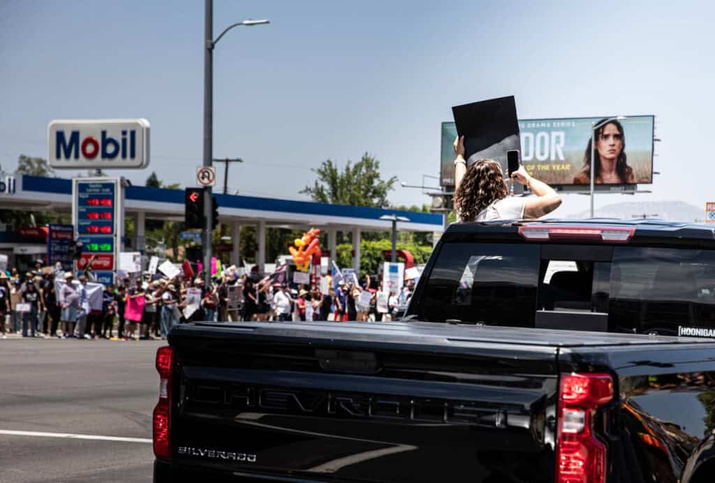 Should influencers get political?? 4 A person stands through the sunroof of a black Chevrolet truck holding a sign at a busy intersection, while protesters with signs, driven by political causes gather near a Mobil gas station in the background.