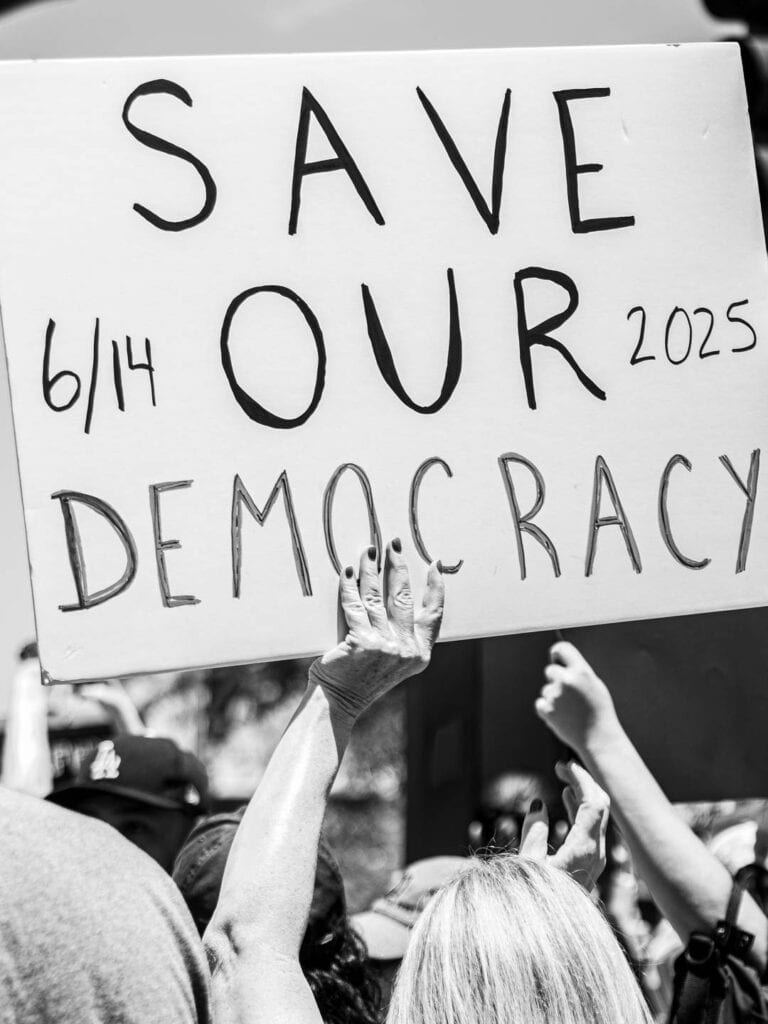 Should influencers get political?? 7 A black-and-white photo of people at a political rally, with a person holding a sign that reads SAVE OUR DEMOCRACY 6/14 2025, surrounded by raised hands—capturing the power of collective action in the age of social media.