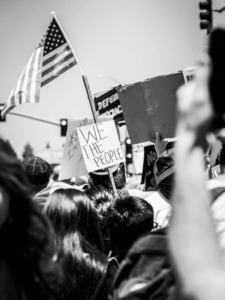 Should influencers get political?? 8 A black-and-white photo of a protest crowd holding signs, including one that reads WE THE PEOPLE and another with an American flag. The energetic, united scene highlights the power of social media in driving political movements under bright daylight.