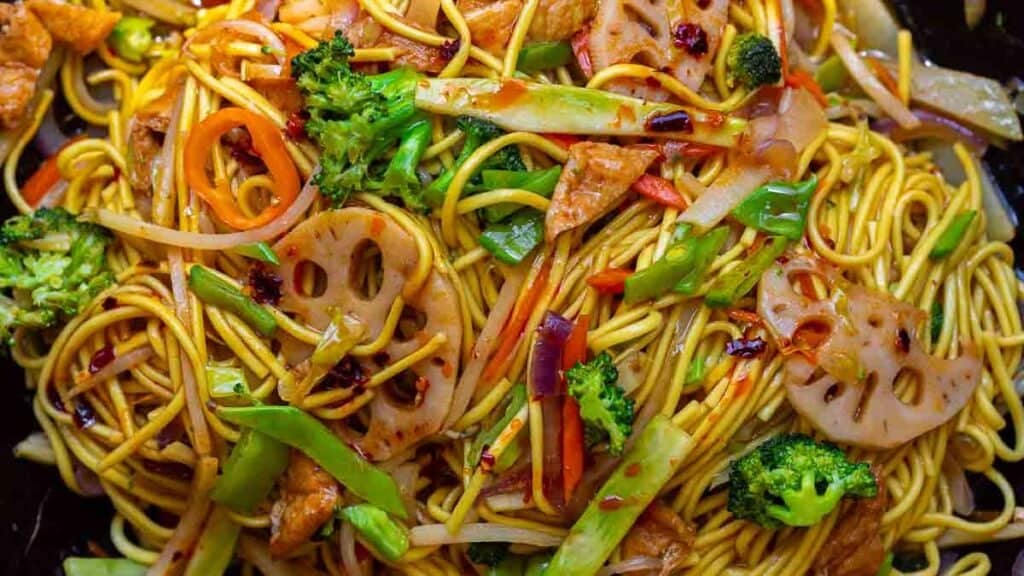 A close-up of colorful stir-fried lo mein vegan noodles mixed with broccoli, carrots, lotus root slices, snow peas, and pieces of tofu, all garnished with chili flakes.