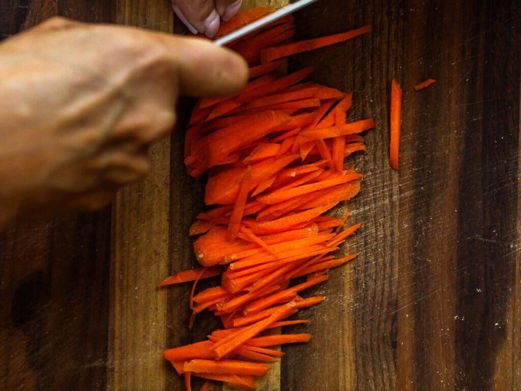 Easy & Healthy Lo Mein Vegan Recipe: Done in Minutes. 23 A hand slices a carrot into thin strips on a wooden cutting board with a kitchen knife.