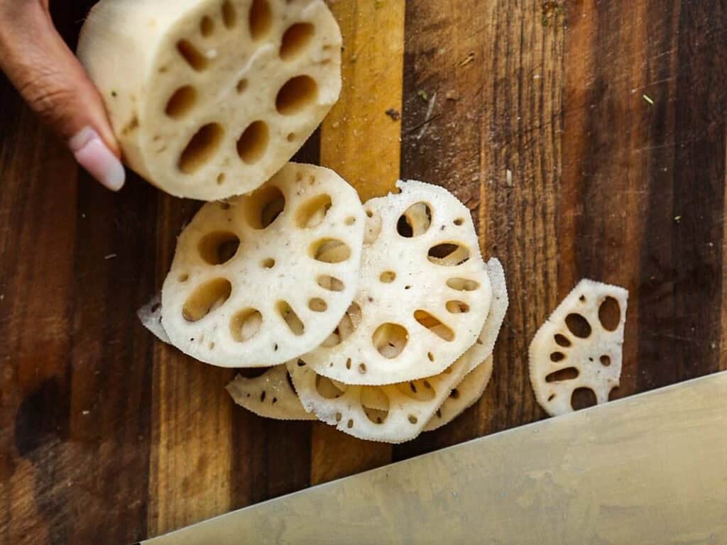 Easy & Healthy Lo Mein Vegan Recipe: Done in Minutes. 24 A hand holds a piece of lotus root above a wooden cutting board, with several round slices of lotus root showing their distinctive holes. A knife is visible at the bottom right.