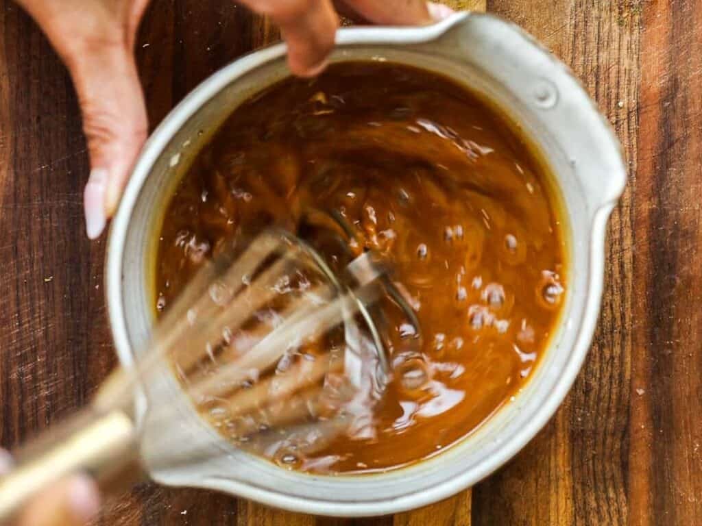 Easy & Healthy Lo Mein Vegan Recipe: Done in Minutes. 28 A person uses a whisk to stir a thick, brown sauce or mixture in a white bowl on a wooden surface.