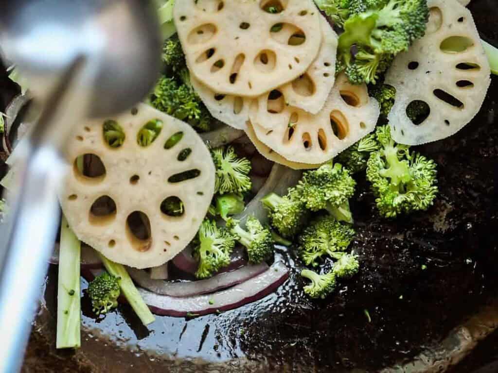 Easy & Healthy Lo Mein Vegan Recipe: Done in Minutes. 30 Sliced lotus root, broccoli florets, and red onion in a pan with oil, being stirred with a metal utensil during cooking.