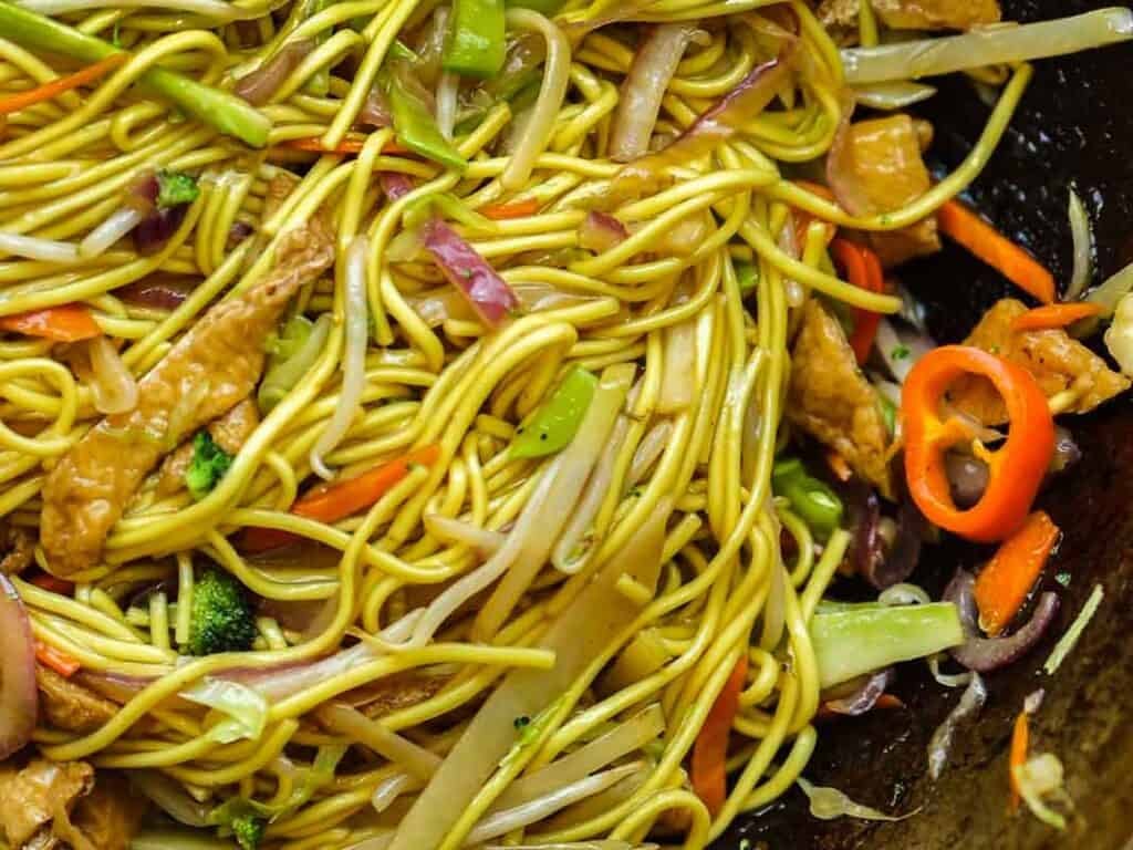 Close-up of vegetable lo mein vegan noodles mixed with sliced red peppers, carrots, broccoli, onions, bean sprouts, and tofu in a wok. The colorful vegetables are intertwined with the yellow noodles.
