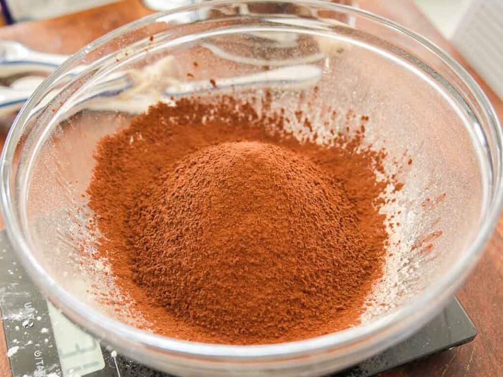 Easy & Delicious Vegan Chocolate Lava Cake Recipe. 37 A glass mixing bowl filled with a mound of sifted cocoa powder sits on a kitchen counter, ready for baking.