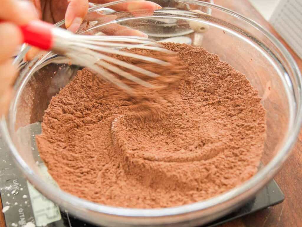 Easy & Delicious Vegan Chocolate Lava Cake Recipe. 38 A hand using a whisk to mix cocoa powder and flour in a clear glass bowl on a kitchen counter.