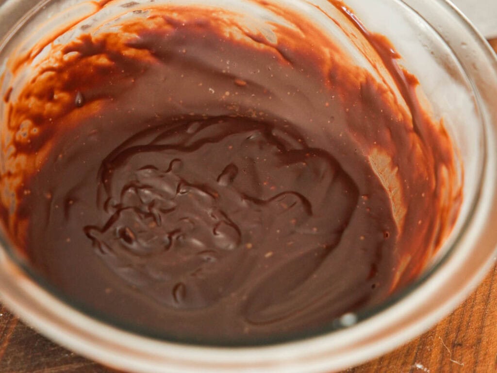 Easy & Delicious Vegan Chocolate Lava Cake Recipe. 32 A glass bowl filled with smooth, glossy hardened chocolate ganache, with some streaks and swirls visible along the edges. The bowl is resting on a wooden surface.