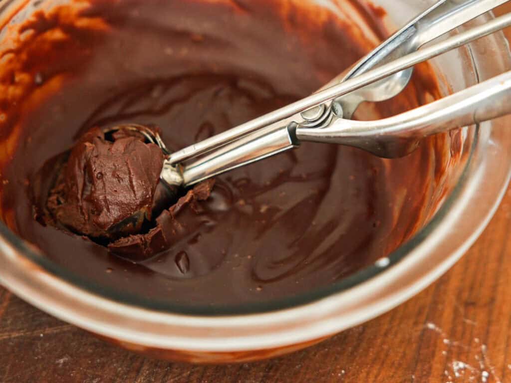 Easy & Delicious Vegan Chocolate Lava Cake Recipe. 33 A metal scoop holds a ball of thick chocolate mixture over a glass bowl filled with melted chocolate on a wooden surface.