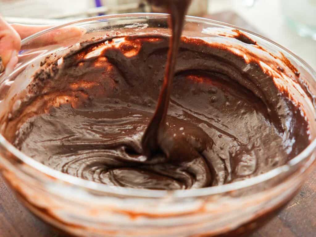 Easy & Delicious Vegan Chocolate Lava Cake Recipe. 39 A close-up of thick chocolate cake batter being mixed in a clear glass bowl, with some batter being lifted by a spoon or spatula.