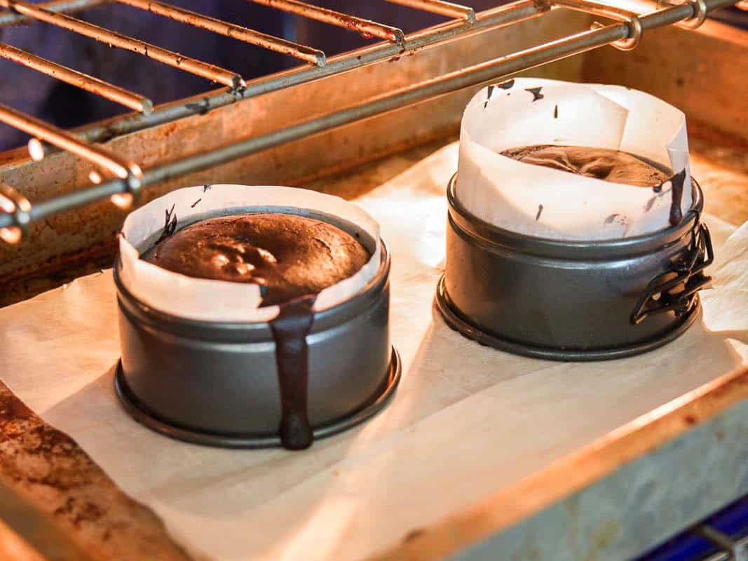 Easy & Delicious Vegan Chocolate Lava Cake Recipe. 25 Two small round cake pans lined with parchment paper, filled with chocolate cake batter, are baking in an oven. One pan has batter overflowing down the side onto the baking sheet below.