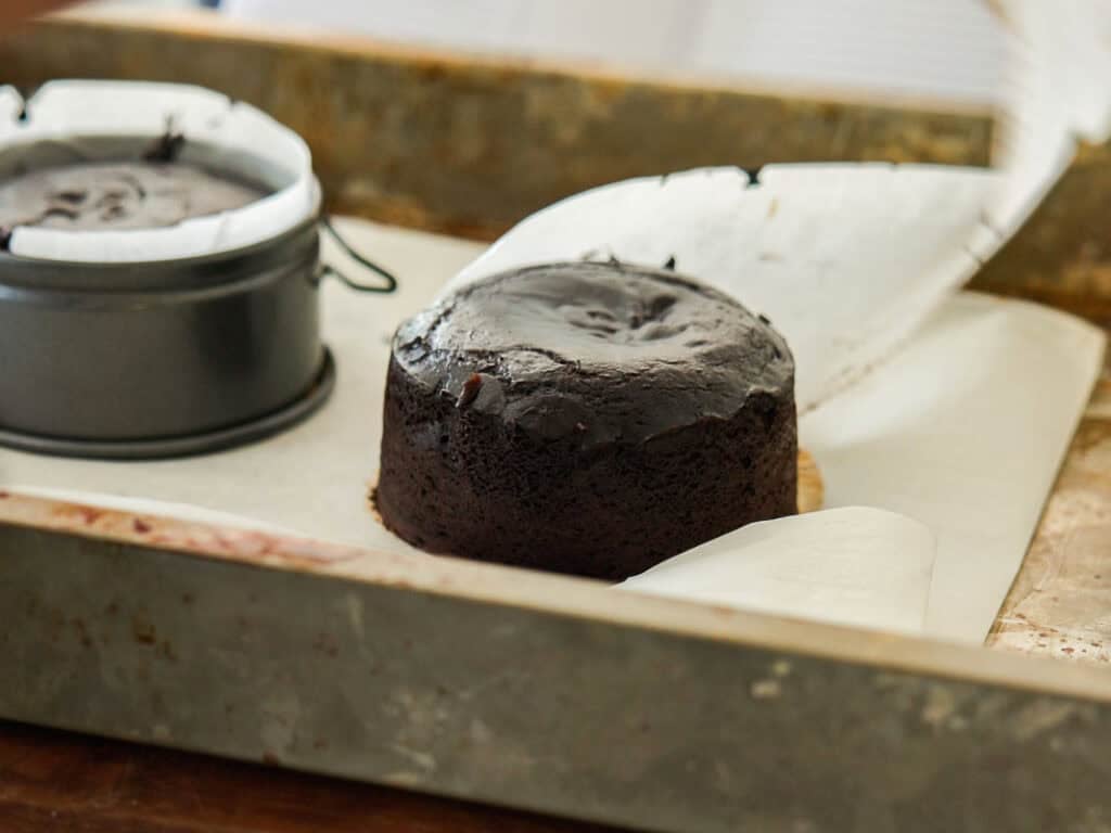 Easy & Delicious Vegan Chocolate Lava Cake Recipe. 24 A freshly baked round chocolate cake sits on parchment paper in a metal baking tray, with another cake still in a round springform pan nearby.
