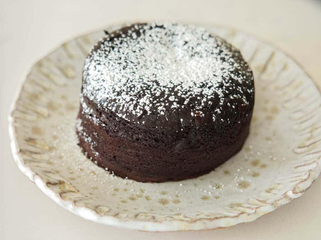 Easy & Delicious Vegan Chocolate Lava Cake Recipe. 44 A round vegan chocolate lava cake topped with powdered sugar sits on a decorative ceramic plate with a scalloped edge.