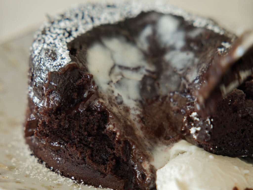 Easy & Delicious Vegan Chocolate Lava Cake Recipe. 19 A close-up of a rich vegan chocolate lava cake with a gooey melted center, topped with a dollop of melted whipped cream and a dusting of powdered sugar. The cake is partially cut, revealing the flowing chocolate inside.