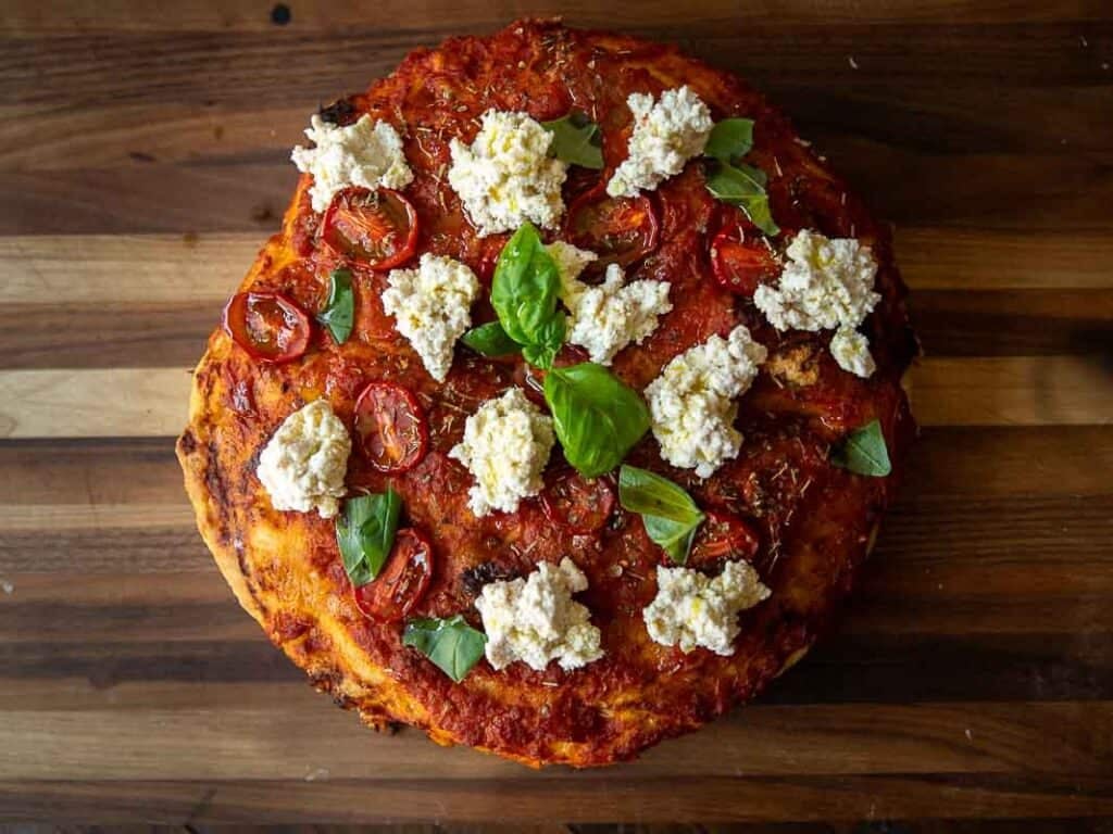 The Perfect Fluffy Focaccia Rossa Recipe - So Easy! 6 A round focaccia rossa topped with tomato sauce, roasted cherry tomatoes, dollops of ricotta cheese, and fresh basil leaves, placed on a wooden surface.