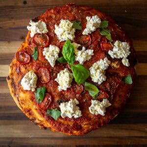 The Perfect Fluffy Focaccia Rossa Recipe - So Easy! 36 A homemade focaccia rossa topped with tomato sauce, roasted tomato slices, dollops of ricotta cheese, and fresh basil leaves, shown on a wooden surface.