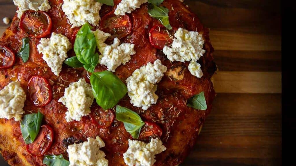 The Perfect Fluffy Focaccia Rossa Recipe - So Easy! 4 A close-up of a rustic focaccia rossa opped with tomato sauce, cherry tomato slices, dollops of ricotta cheese, fresh basil leaves, and herbs on a wooden surface.