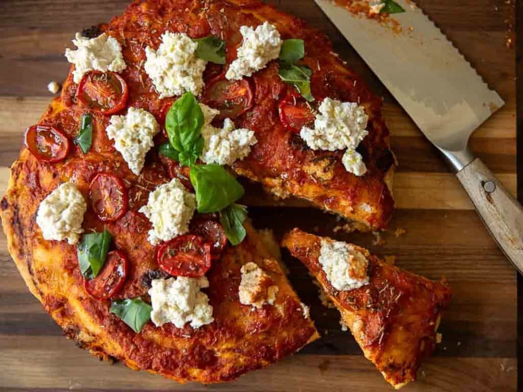 The Perfect Fluffy Focaccia Rossa Recipe - So Easy! 5 A focaccia rossa topped with tomato sauce, fresh basil leaves, sliced cherry tomatoes, and dollops of vegan ricotta cheese on a wooden board. A slice is being separated with a large knife.