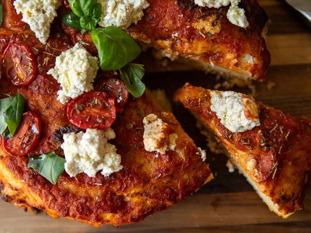 The Perfect Fluffy Focaccia Rossa Recipe - So Easy! 8 A close-up of a focaccia rossa topped with tomato sauce, roasted cherry tomatoes, dollops of vegan ricotta, and fresh basil leaves on a wooden surface. One slice is slightly pulled away from the pizza.