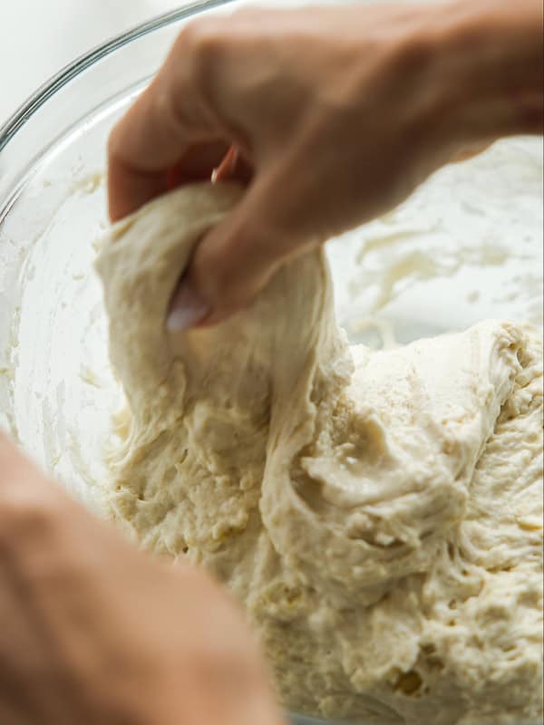 The Perfect Fluffy Focaccia Rossa Recipe - So Easy! 12 Close-up of hands stretching and folding soft bread dough in a glass bowl. The dough appears sticky and airy, indicating it is in the mixing or kneading stage of preparation.