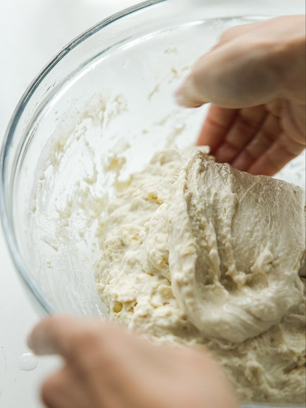 The Perfect Fluffy Focaccia Rossa Recipe - So Easy! 13 Close-up of hands kneading or folding sticky bread dough in a clear glass bowl. The dough appears soft and elastic, and the background is bright and out of focus.