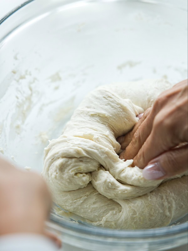 The Perfect Fluffy Focaccia Rossa Recipe - So Easy! 14 A close-up of hands kneading dough in a clear glass bowl, with soft, stretchy dough being pressed and folded.