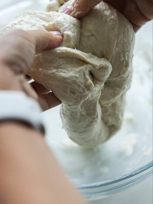 The Perfect Fluffy Focaccia Rossa Recipe - So Easy! 15 Close-up of hands stretching and folding sticky bread dough in a glass bowl, preparing it for baking.
